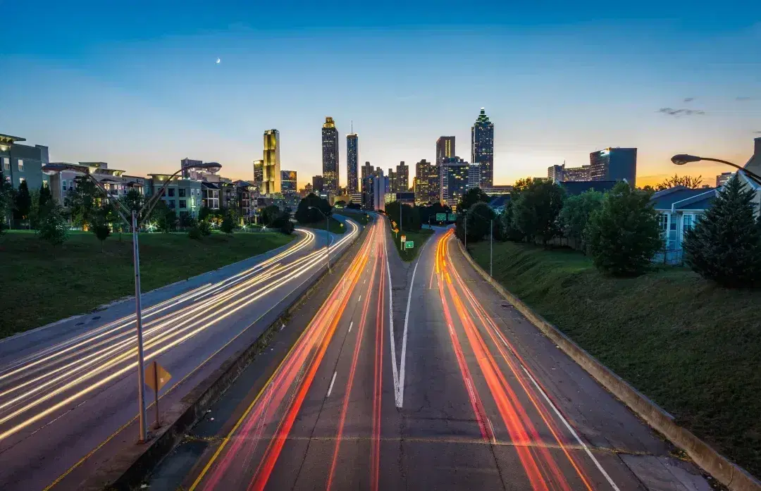 Headlight trails on a highway leading into a city at dusk.